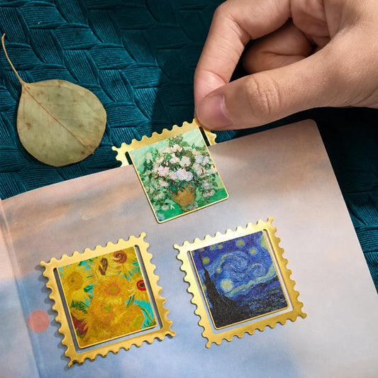 Trois marque-pages timbres dorés (Vase de roses, Tournesols, Nuit étoilée) posés sur un livre ouvert, une main en saisit un